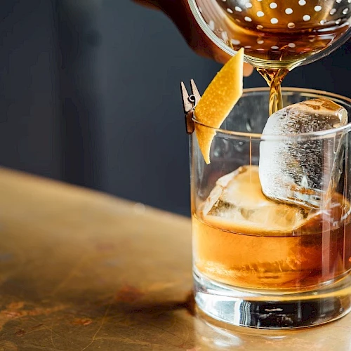 A short, amber cocktail being poured into a short glass with a large ice cube and a twist of orange peel garnish.