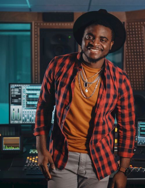 A smiling man in a red plaid shirt and orange tee stands in a music studio with mixing consoles and large speakers behind him, wearing a hat.