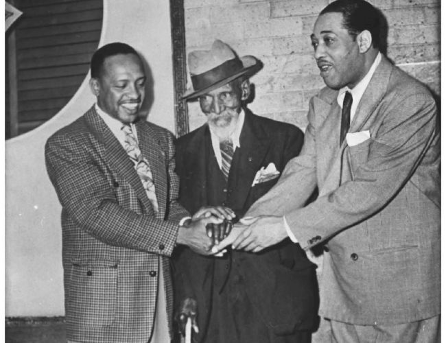 Three men in suits are shaking hands in a black-and-white photo. One wears a hat and uses a cane, standing in front of a brick wall.