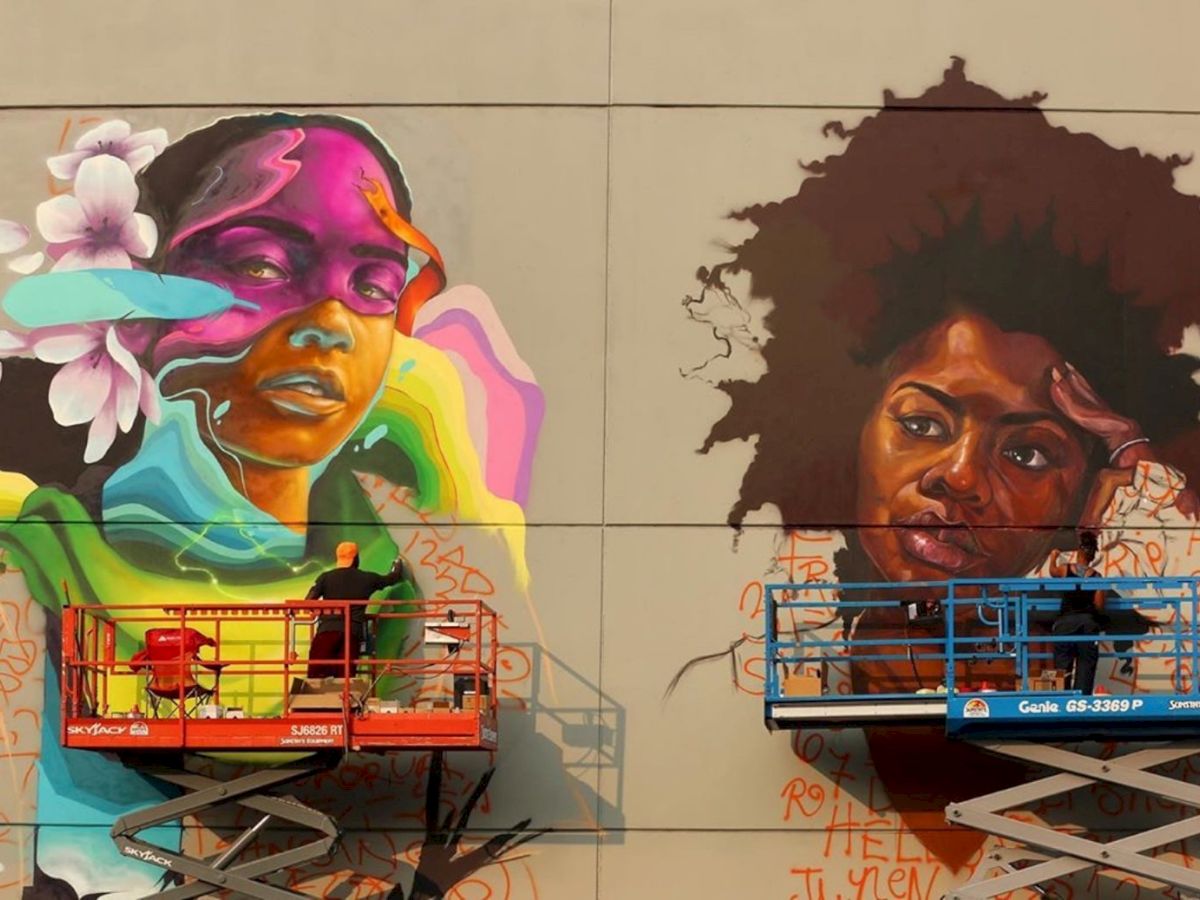 Two artists work on vibrant mural panels: left side shows a colorful portrait with flowers and bold shapes, right side features a woman with natural hair.